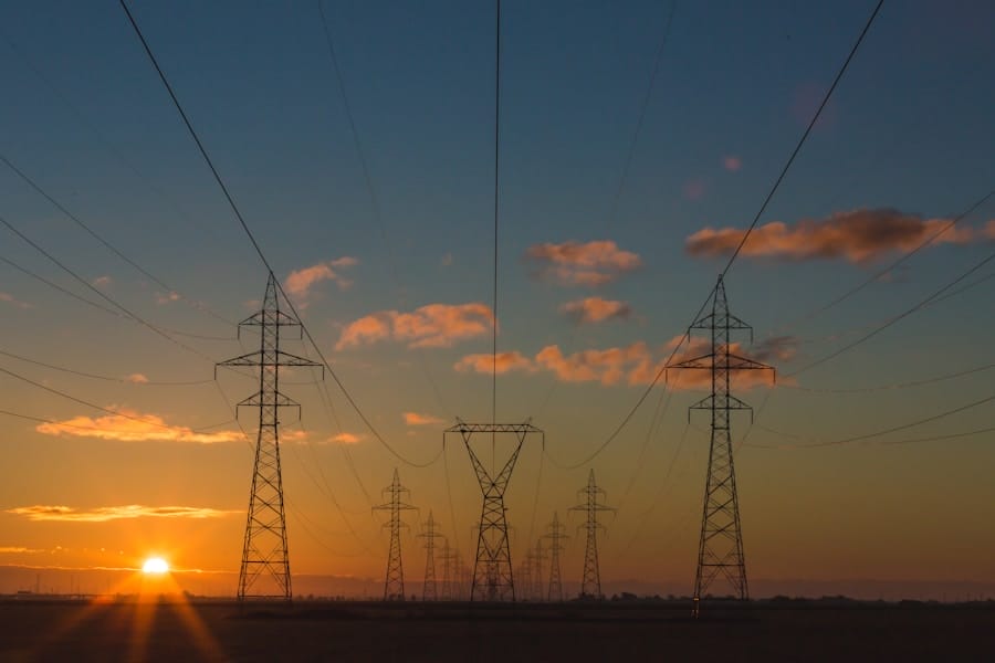 Electric power transmission towers and lines stretch across a landscape at sunset, with the sun low on the horizon and scattered clouds in the colorful sky.
