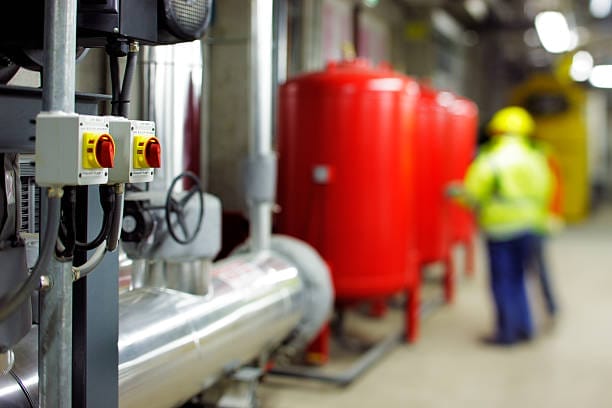 Industrial setting with large red tanks, control valves, and piping in the foreground; two workers in safety gear and yellow helmets stand in the blurred background—ideal for a Fire Safety Audit to ensure all fire safety measures are properly maintained.