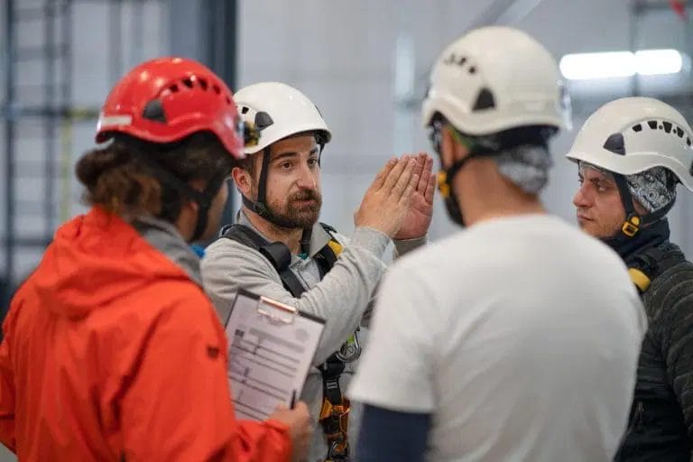 A man in a safety harness and helmet gestures while discussing a safety audit with three others in helmets and work gear; one person holds a clipboard.