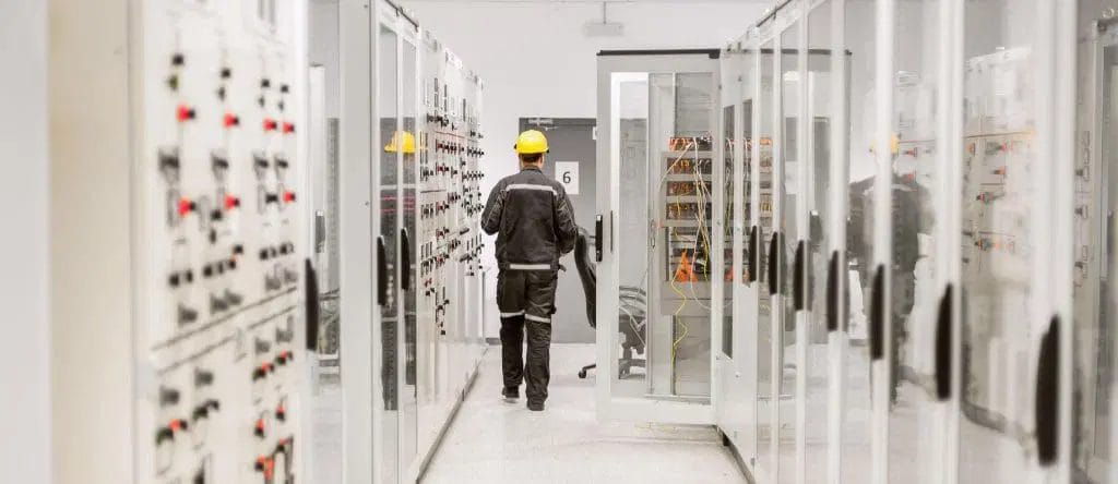A worker in a yellow hard hat and protective clothing walks through a corridor lined with electrical control panels, performing Power Quality Analysis in an industrial facility.