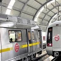 Two silver Metro Rail trains with yellow stripes and red logos are stopped at an indoor, arched-roof station platform in Delhi NCR, reflecting a high standard of public transport safety and compliance.