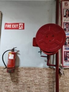 A red fire extinguisher and fire hose reel are mounted on a tiled wall under a red Fire Exit sign, beside photo posters—an ideal setup to feature in a fire audit for safety compliance.