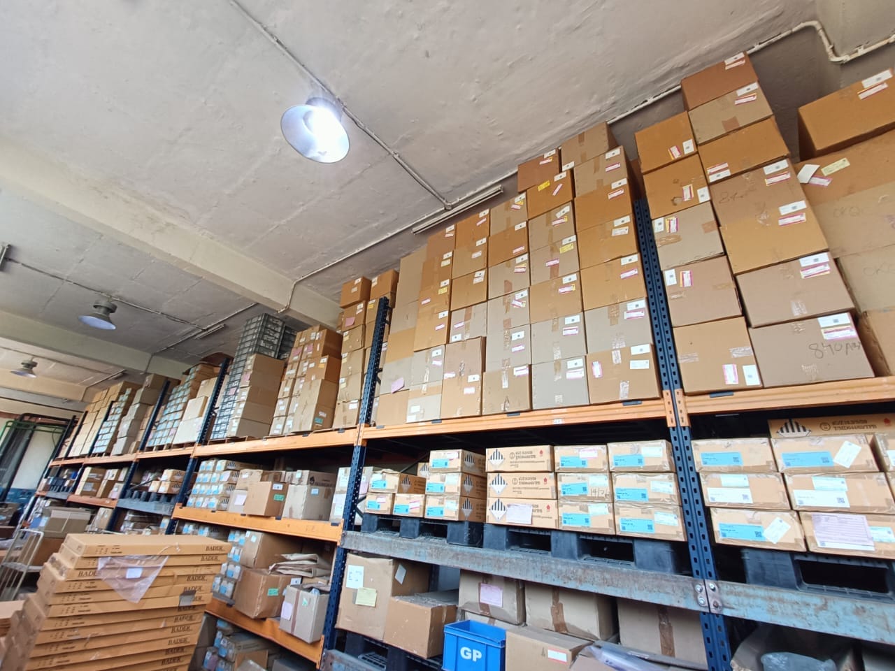 Rows of metal shelves filled with stacks of brown cardboard boxes are organized in a warehouse with a concrete ceiling and fluorescent lighting, ready for inventory or storage items and an upcoming fire safety audit to ensure compliance.