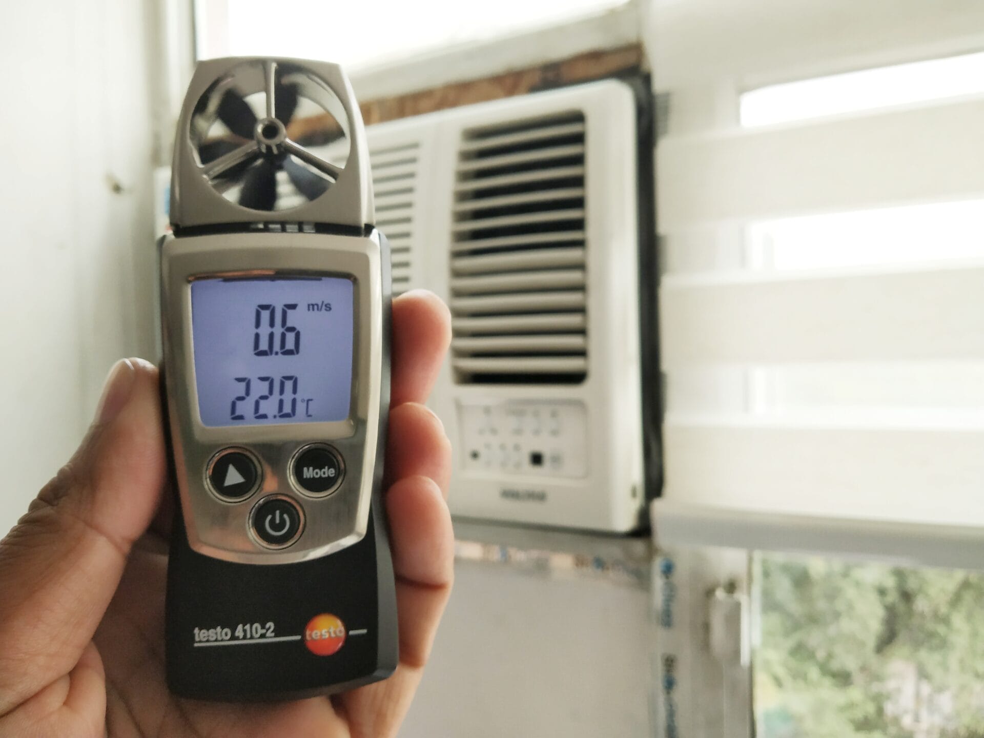 A hand holds an anemometer showing 0.6 m/s and 22.0°C in front of a window air conditioner unit, capturing airflow data as part of an energy audit with partially open blinds in the background.