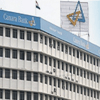 A multi-story white building with Canara Bank signage and logo on the facade and rooftop, along with the Indian flag flying above.