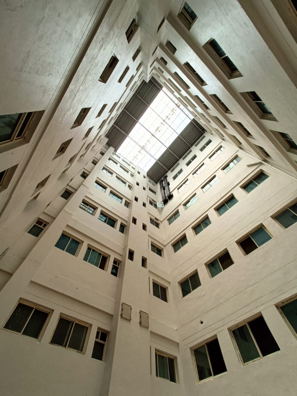 View looking up from the bottom of a tall, white-walled, multi-story building with many windows, leading to a glass ceiling that lets in natural light.