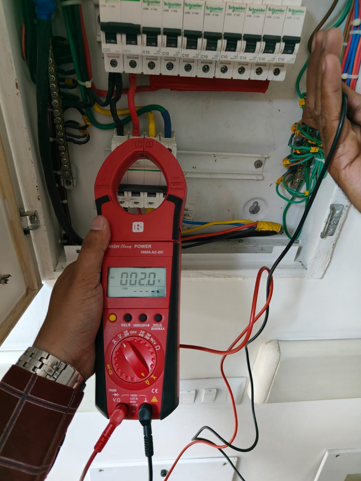 A person uses a red digital clamp meter to measure electrical current in a circuit breaker panel, with the device display showing “0.20” and various colored wires visible inside the panel.