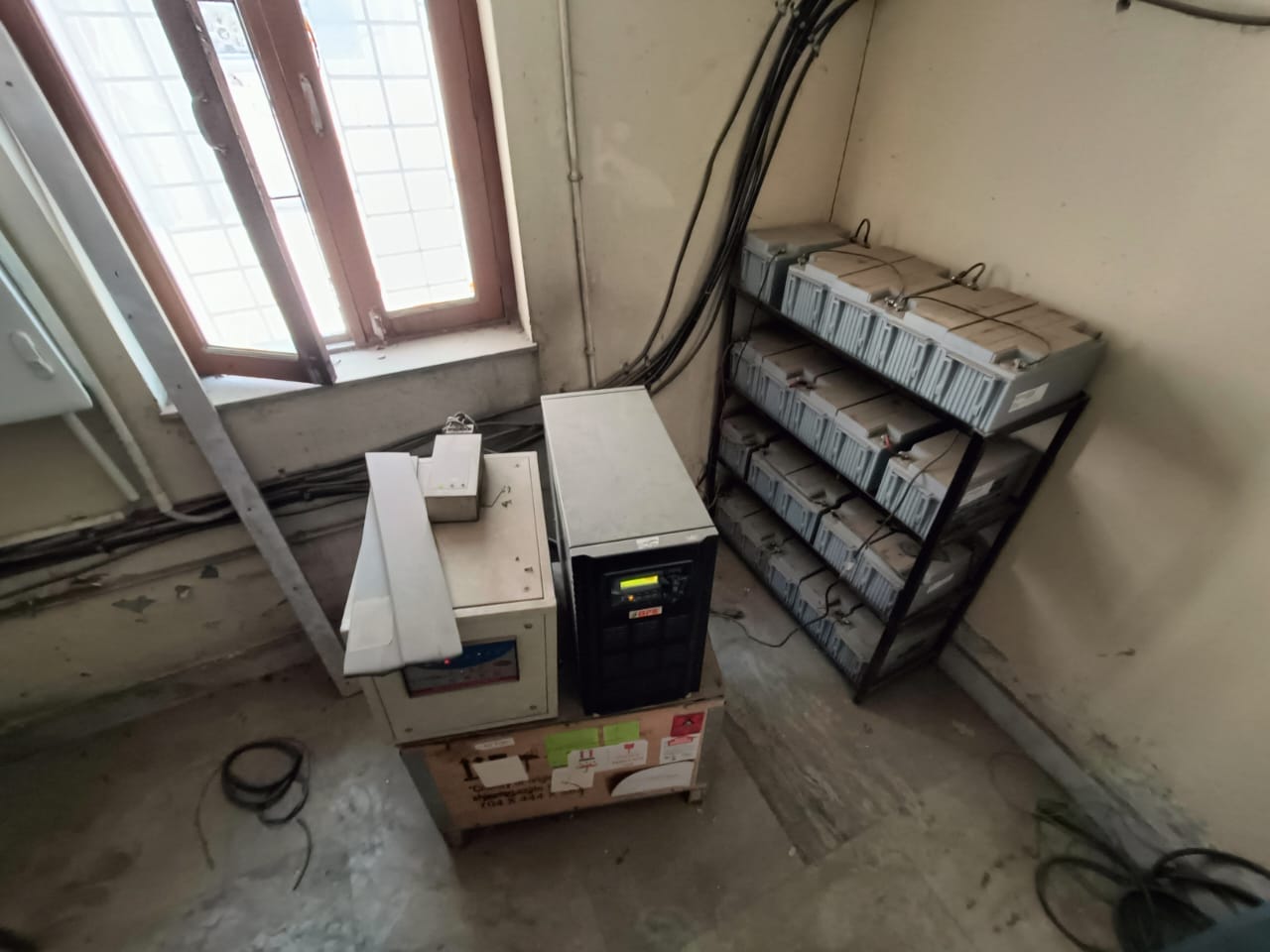 A small room with electronic equipment, including two machines on a box and a metal shelf holding multiple grey battery packs. Cables run along the walls, and sunlight comes through a window with bars. The floor is bare concrete.