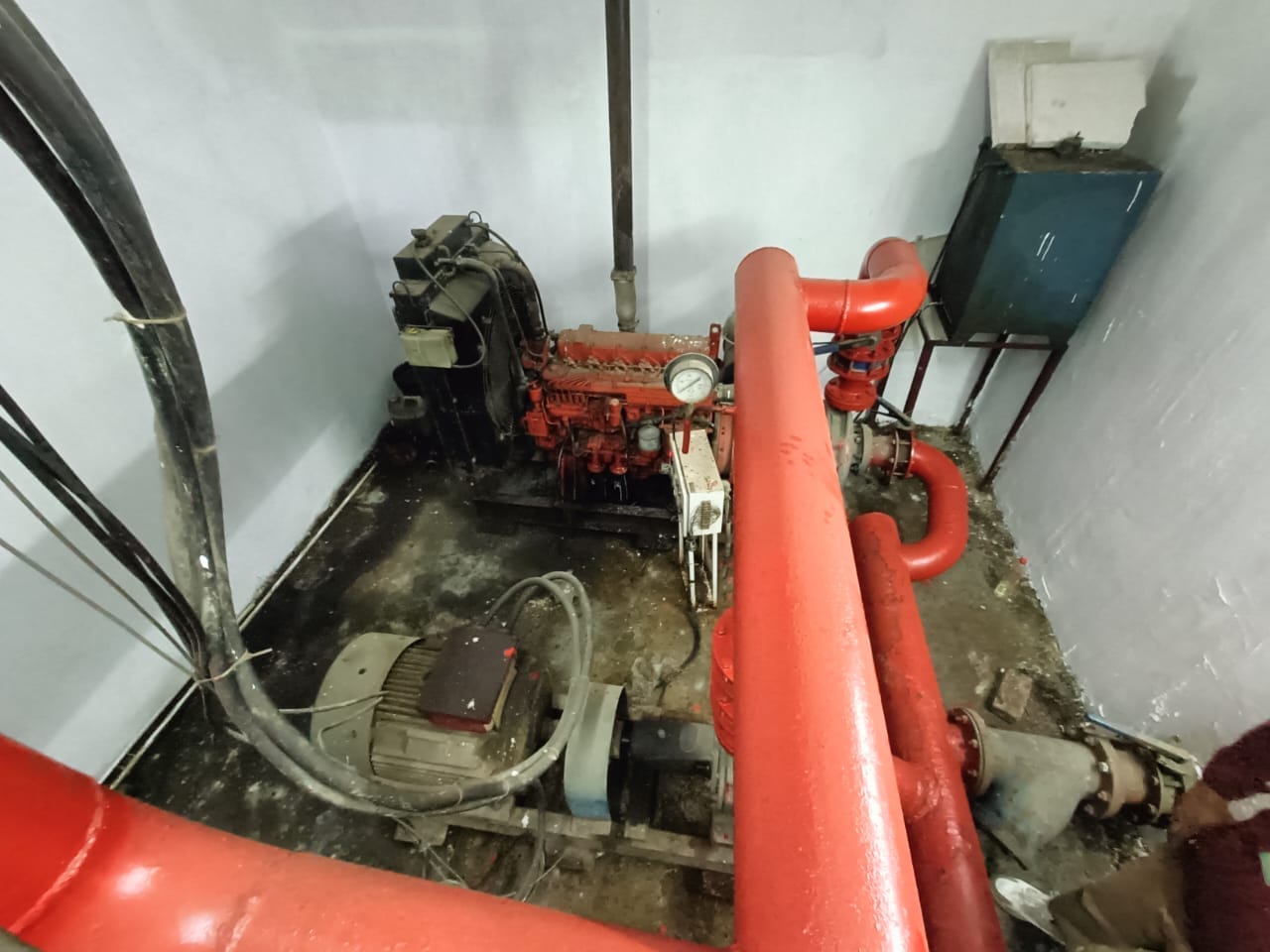 A room with industrial equipment, including a red pipe system, a pump, and a control panel mounted on the wall. The floor appears worn and the setting looks like a utility or mechanical room.
