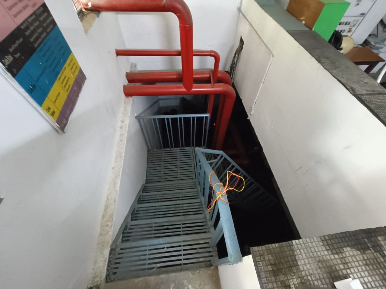 A metal staircase leads downwards in a narrow indoor space with white walls and red pipes overhead. An orange cable is tied to the blue railing, and nearby are colorful signs and office furniture.
