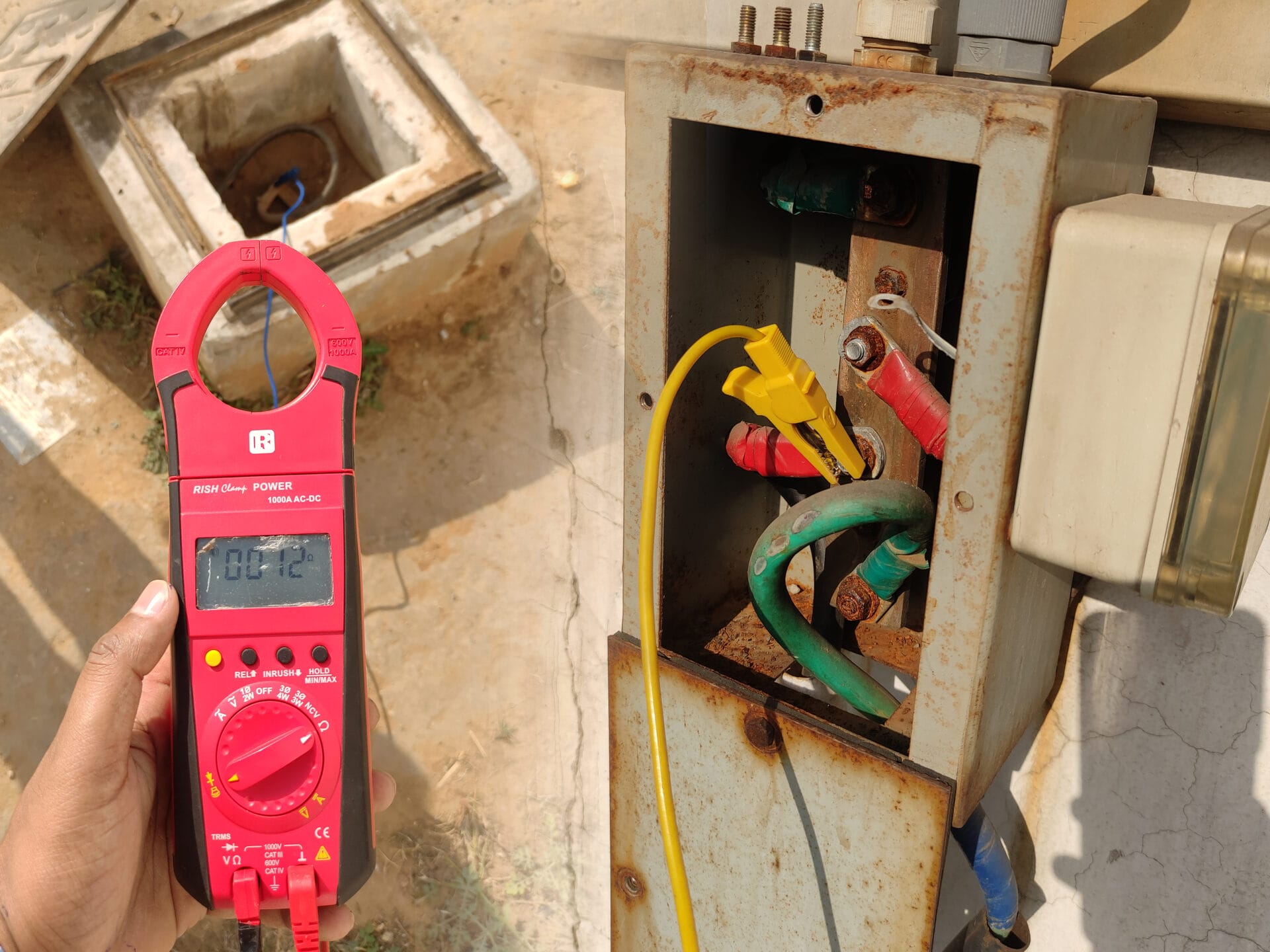 A person holds a red digital clamp meter displaying 12 volts near an open, rusty electrical panel with colored wires connected inside, outdoors on a sunny day.
