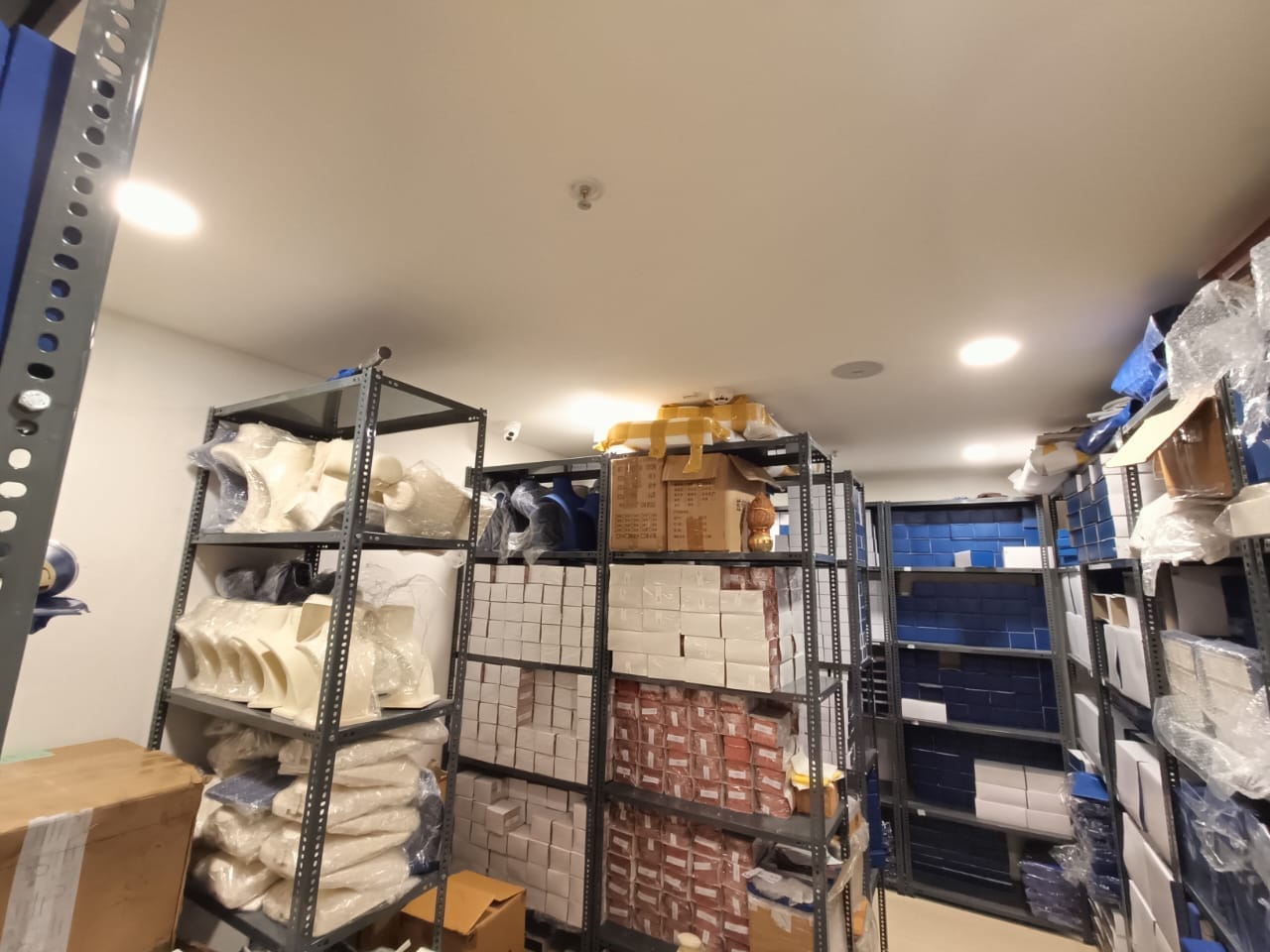 Metal shelves filled with boxes and various packaged items line the walls of a well-lit storage room with a light-colored ceiling and floor.