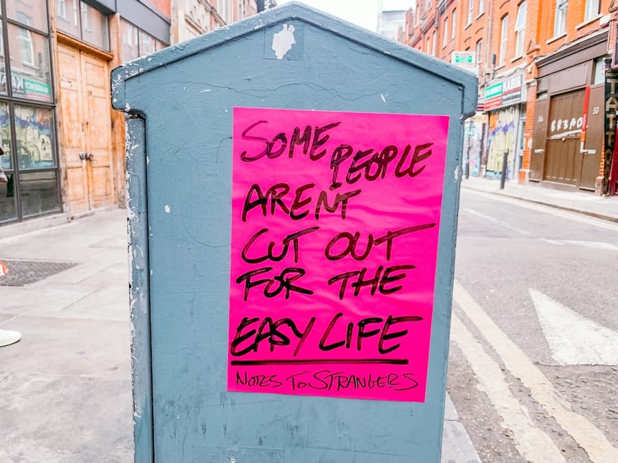 A bright pink poster on a gray utility box reads, Some people arent cut out for the easy life. Notes to strangers, on a city street with brick buildings in the background.