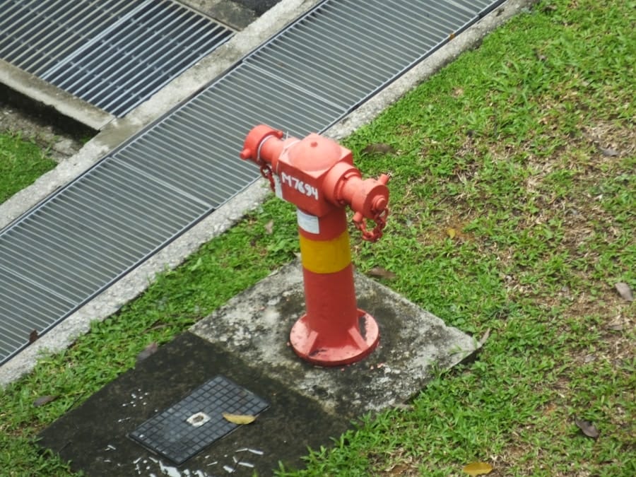 A red fire hydrant with a yellow band is installed on a concrete base next to a grassy area and near metal grates embedded in the ground.