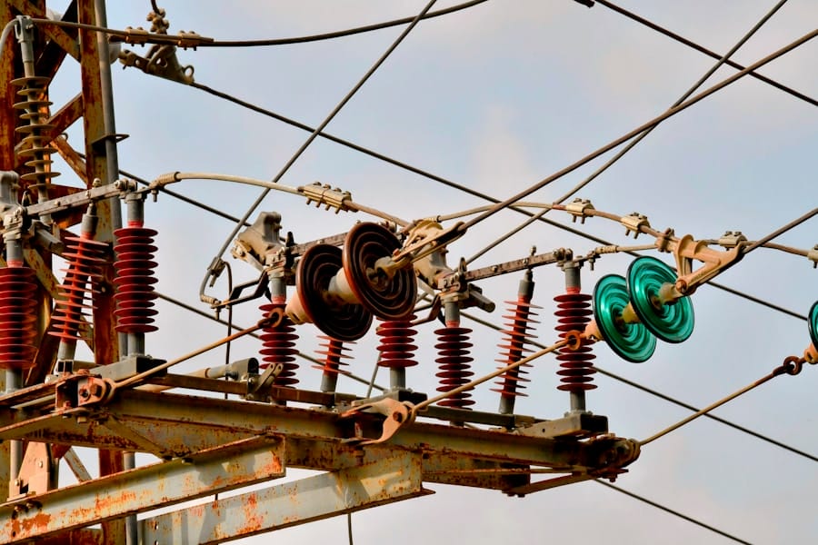 Close-up of high-voltage power lines and electrical insulators attached to a metal structure, with some coils and ceramic disks visible against a blue sky.