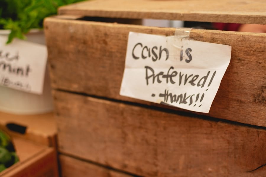 A handwritten sign taped to a wooden crate reads Cash is preferred! -thanks!! with some greenery and a white container in the background.
