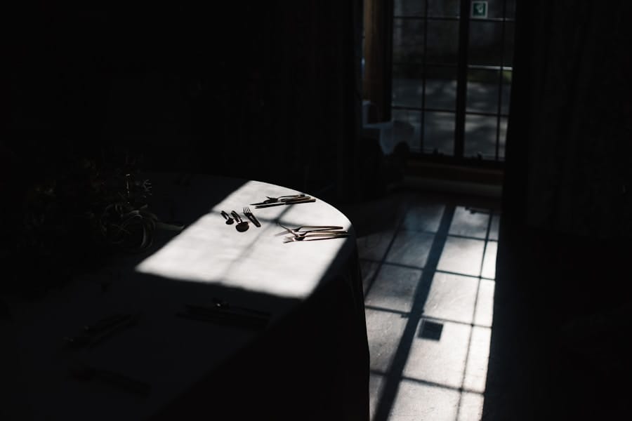 A round table with cutlery is partially illuminated by sunlight streaming through a window, casting shadows across the floor in an otherwise dimly lit room.