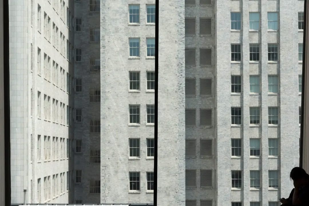 A person stands near a window in silhouette, looking out at a tall, gray brick building with many windows. The scene is divided by a vertical window frame.