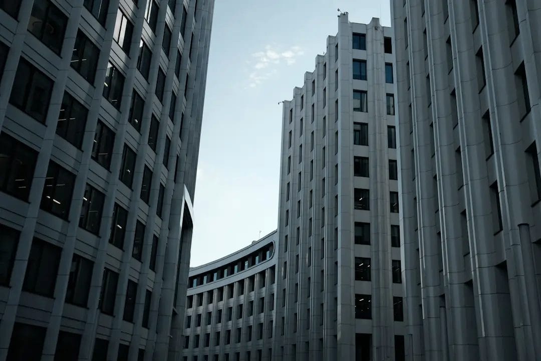 Tall, modern office buildings with many windows create a narrow corridor, leading to a curved building in the background, against a partly cloudy sky.